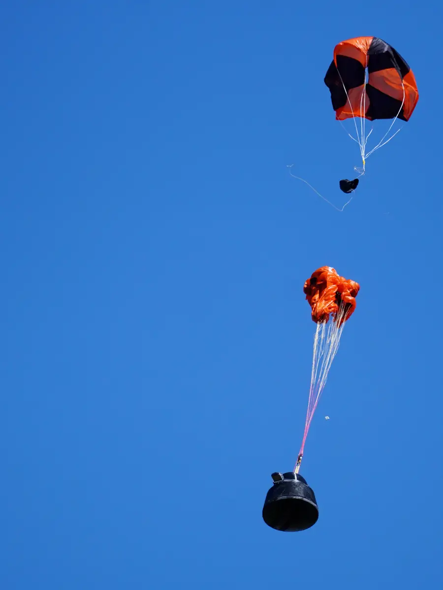 A pod descending under two drogue parachutes against a blue sky
